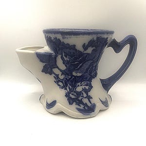 Op de afbeelding: Een vintage scheerbeker met een blauw en wit bloemmotief. De keramische beker heeft een unieke vorm met een ingebouwde zeepbak en een hartvormig handvat. Een klassiek stuk voor traditioneel scheren.