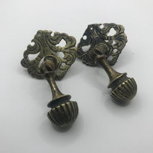 May include: Two antique brass drawer pulls with ornate, openwork designs. Each pull features a decorative backplate and a ribbed, rounded handle. The pulls are a warm, aged brass colour, suggesting vintage or retro style.