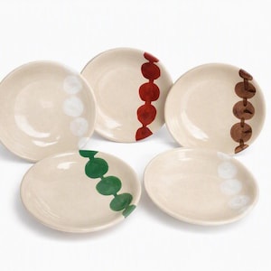 May include: Five round ceramic plates with a cream-colored base. Each plate features a single vertical line of five painted circles in different colors, including white, red, brown, and green, arranged in a row across the surface of the dish.