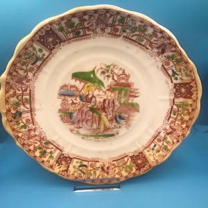 May include: Antique ceramic plate with a scalloped edge. The plate features a central scene of figures under a green umbrella, surrounded by a decorative border in shades of brown, pink, and green. The plate has a diameter of approximately 9 inches.