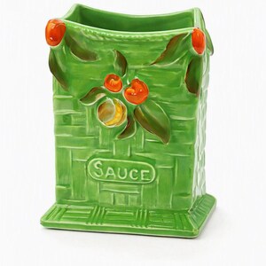 Pode incluir: Um suporte de utensílios de cerâmica verde com textura de cestaria. A frente apresenta frutos laranja e folhas verdes em relevo, com a palavra SAUCE impressa numa moldura oval elevada. O recipiente tem uma base quadrada e topo aberto.