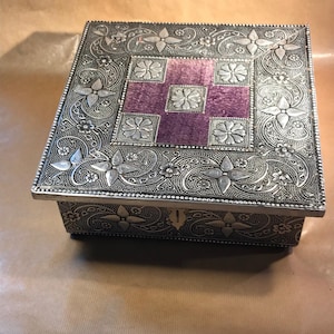 Könnte beinhalten: Eine silberne Metallbox mit einem verzierten Blumendesign und einem violetten Samt-Innenraum. Die Box hat einen Klappdeckel und einen kleinen Riegel.