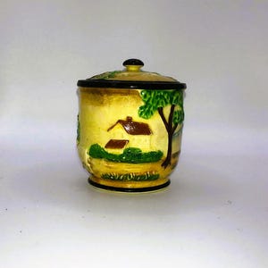 Pode incluir: Um pote de cerâmica com tampa, com um design de paisagem rural. O pote é principalmente amarelo com uma borda preta e um botão preto na tampa. A paisagem inclui uma casa, uma árvore e grama verde.