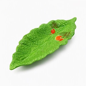 Puede incluir: Un plato de servicio de cerámica con forma de hoja verde texturizada y superficie irregular. La hoja presenta dos pequeños detalles pintados en color naranja brillante en la parte superior derecha, que imitan la apariencia de frutos o bayas.