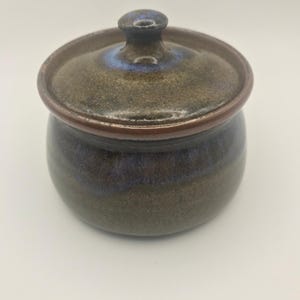 Op de afbeelding: Een kleine, ronde keramische pot met een deksel. De pot is geglazuurd in tinten bruin, blauw en groen. Het deksel heeft een kleine knop bovenop. De pot is waarschijnlijk handgemaakt en heeft een rustieke uitstraling.