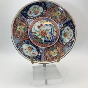 May include: A round ceramic plate featuring a central circular medallion with floral and bamboo motifs, surrounded by a complex border of geometric patterns, dark blue panels, and cloud-shaped vignettes containing colorful flowers.