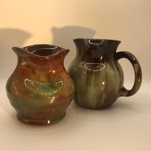 May include: Two ceramic jugs with a glossy finish. One is a small vase-like jug with a flared rim, and the other is a larger jug with a handle. The jugs have a blend of brown, green, and orange hues.