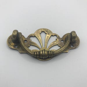 Könnte beinhalten: Antiker Messing-Schubladengriff mit fächerförmigem Design. Der Griff ist gebogen und an einer dekorativen Rückplatte mit zwei Schraubenlöchern auf jeder Seite befestigt. Die Hardware hat eine Vintage-Ästhetik.