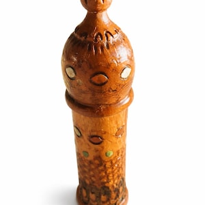 Puede incluir: Un objeto cilíndrico de madera con una parte superior redondeada, que presenta una textura tallada y detalles circulares incrustados en tonos blanco, verde y marrón. La superficie muestra un acabado de veta natural con patrones grabados.