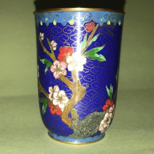 Puede incluir: Un pequeño jarrón de cloisonné azul y dorado con un diseño floral. El jarrón está decorado con una rama de flores blancas y rosas con hojas verdes.