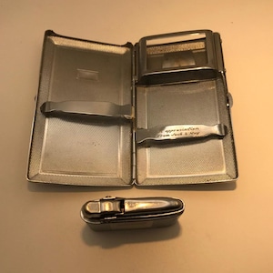 May include: An open, silver-toned cigarette case with a textured interior and a separate compartment. The case has a curved metal holder and the inscription "In appreciation From Jack & May". A small, silver-toned lighter is below the case.