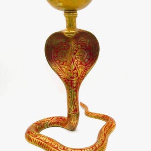 May include: A brass cobra-shaped candle holder with red and gold accents. The candle holder has a detailed design and a decorative bowl at the top. The cobra's body is coiled, and the head is raised.