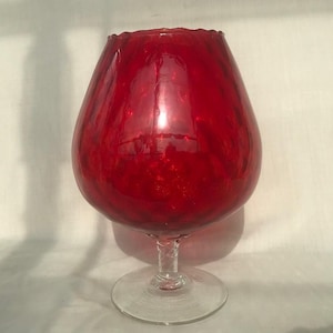 Könnte beinhalten: Ein großes, rotes Brandyglas mit breiter Schale und klarem, gedrehtem Stiel. Der Rand der Schale hat einen gewellten Rand. Das Glas hat eine glänzende Oberfläche und steht auf einem klaren, runden Fuß.