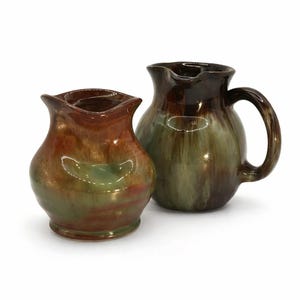 May include: Two ceramic jugs featuring a glossy, multi-coloured glaze in shades of deep brown, forest green, and warm amber. The smaller jug has a wide, flared rim, while the larger jug includes a curved handle and a pouring spout.