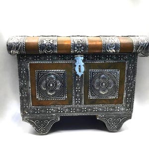 May include: A decorative chest with intricate silver and copper metalwork. The chest features floral designs and a rectangular shape with a curved top. The front has a central lock and decorative panels. The chest is supported by four ornate feet.
