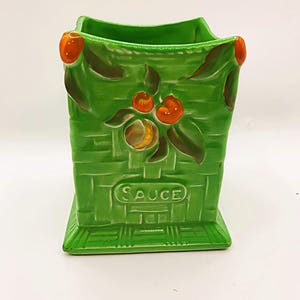 May include: A green ceramic sauce holder with a basket weave design. The holder features orange fruit and leaf accents. The word "SAUCE" is embossed on the front. The holder has a rectangular base and a square top.