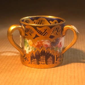 Puede incluir: Una taza pequeña de porcelana con dos asas, adornada con ribetes dorados. La taza está decorada con un patrón azul y dorado y diseños florales.