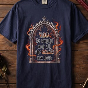 May include: A navy blue short-sleeve t-shirt featuring a graphic of a gothic archway surrounded by orange flames. The text inside the archway reads, "Hell is empty and all the devils are here," written in a stylised, dark, gothic-inspired font.