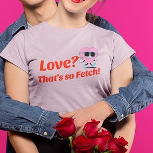 Może przedstawiać: Para w pozie do zdjęcia na tle różowego tła. Kobieta ma na sobie jasnolilawy t-shirt z napisem "Love? That's so Fetch!" i dżinsową kurtkę. Mężczyzna ma na sobie dżinsową kurtkę. Trzymają bukiet czerwonych róż.