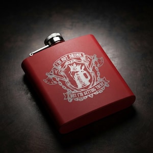 May include: A matte red metal flask featuring a silver-toned screw-top cap. The front displays a white engraved graphic of a beer mug with a crown, flanked by two crossed axes and chains, with the text I'M NOT DRUNK BUT I'M GETTING THERE.