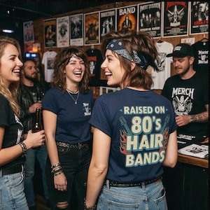May include: A navy blue t-shirt featuring the text "RAISED ON 80's HAIR BANDS" in a retro font, flanked by two electric guitar graphics with orange and red flames. The shirt is worn by a person with a dark blue patterned bandana in their hair.