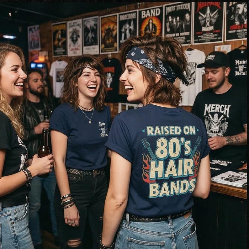 May include: A navy blue t-shirt featuring the text "RAISED ON 80's HAIR BANDS" in a retro font, flanked by two electric guitar graphics with orange and red flames. The shirt is worn by a person with a dark blue patterned bandana in their hair.