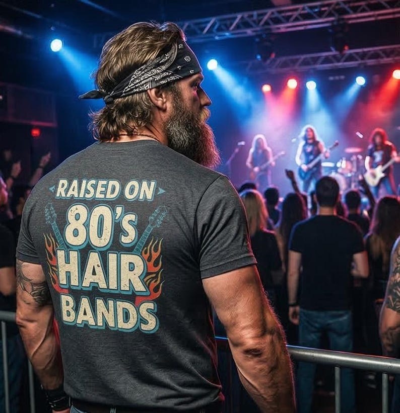 May include: A charcoal gray short-sleeve t-shirt featuring a graphic on the back with the text RAISED ON 80's HAIR BANDS in light blue and white, flanked by two electric guitar illustrations and orange flame accents in a distressed style.