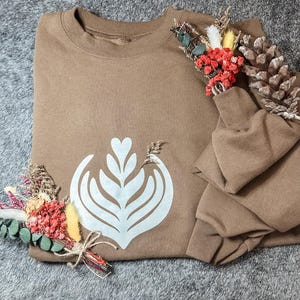 May include: A brown sweatshirt with a white latte art design. The folded sweatshirt is decorated with dried flowers and a pinecone. The sleeves are folded on top of the sweatshirt.