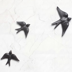 Puede incluir: Tres decoraciones de pared con forma de golondrina en color negro mate dispuestas en formación de vuelo. Cada ave presenta texturas detalladas en las plumas de las alas y una cola bifurcada, capturando a las aves en pleno vuelo.