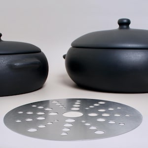 Könnte beinhalten: Ein Set aus schwarzen Keramik-Kochtöpfen mit Deckeln und einem silbernen Dampfeinsatz aus Metall mit unterschiedlich großen Löchern. Die Töpfe haben abgerundete Körper und kleine Griffe. Der Dampfeinsatz ist kreisförmig.