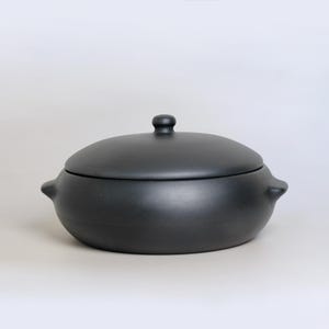 Peut inclure: Un plat à casserole en céramique noire mate avec un couvercle. Le plat a une forme arrondie, deux petites poignées sur les côtés et un petit bouton sur le couvercle. Design simple et moderne.