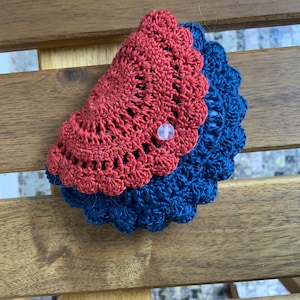 May include: A handmade crochet wallet in two colors, rust and navy blue. The wallet has a scalloped edge and is secured with a clear button. The openwork design is visible, showcasing the intricate crochet pattern. The wallet is resting on a wooden surface.