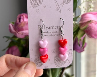Op de afbeelding: Een paar oorhangers met drie gelaagde hartcharms. De harten zijn rood, roze en lichtroze. De oorhangers bevinden zich op een witte kaart met de tekst "Rhyarncraft @rhyarncraft rhyarncraft.etsy.com".