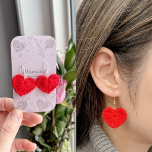 May include: Red crocheted heart earrings on a white card with the text "RHyarncraft" and a crochet hook. The earrings are also shown on a person's ear.