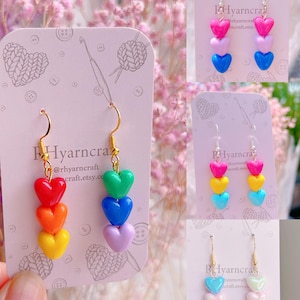 Puede incluir: Pendientes con dijes en forma de corazón en colores del arcoíris, con ganchos dorados. Los pendientes presentan tres corazones apilados en rojo, naranja, amarillo, verde, azul y morado. Otros pendientes en la imagen tienen corazones rosas, amarillos, azules e iridiscentes. El texto "Hyarncra" está presente.