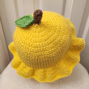 May include: A yellow crocheted hat shaped like a lemon with a green leaf and brown stem on top.