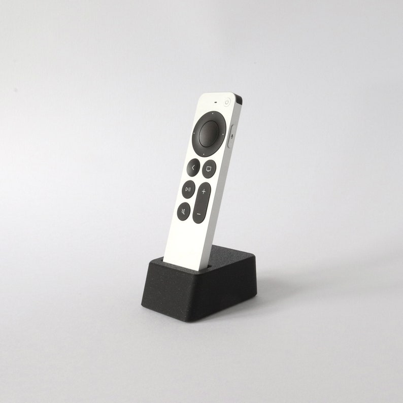 May include: A white remote control with a circular button and several other buttons, including a play/pause button, a plus button, and a minus button. The remote is sitting on a black charging stand.