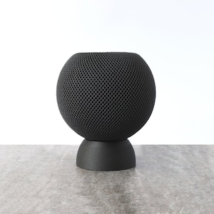 Könnte beinhalten: Ein schwarzer, kugelförmiger Lautsprecher mit einem gitterartigen Äußeren, der auf einem passenden dunkelgrauen Sockel steht. Der Lautsprecher steht auf einer hellgrauen, strukturierten Oberfläche vor einem weißen Hintergrund. Modernes, minimalistisches Design.