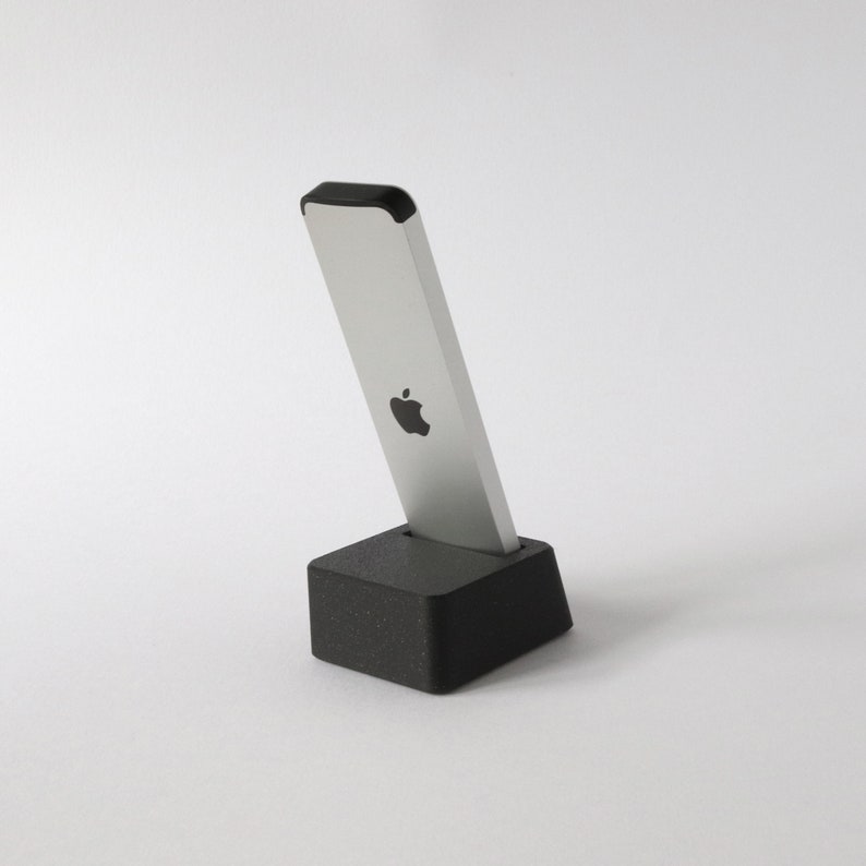 May include: A silver Apple remote control with a black top and an Apple logo on the front, sitting on a black charging dock.