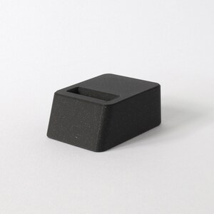 May include: A black, textured, rectangular phone stand with a slot for charging.
