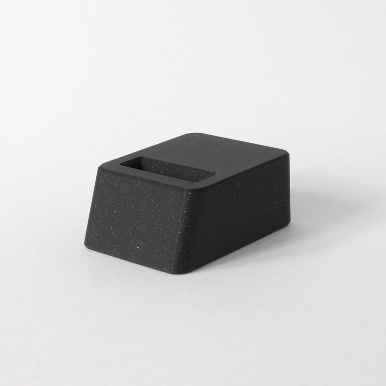 May include: A black, textured, rectangular phone stand with a slot for charging.