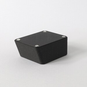 May include: Black square-shaped box with a textured surface and four silver dots on the top.