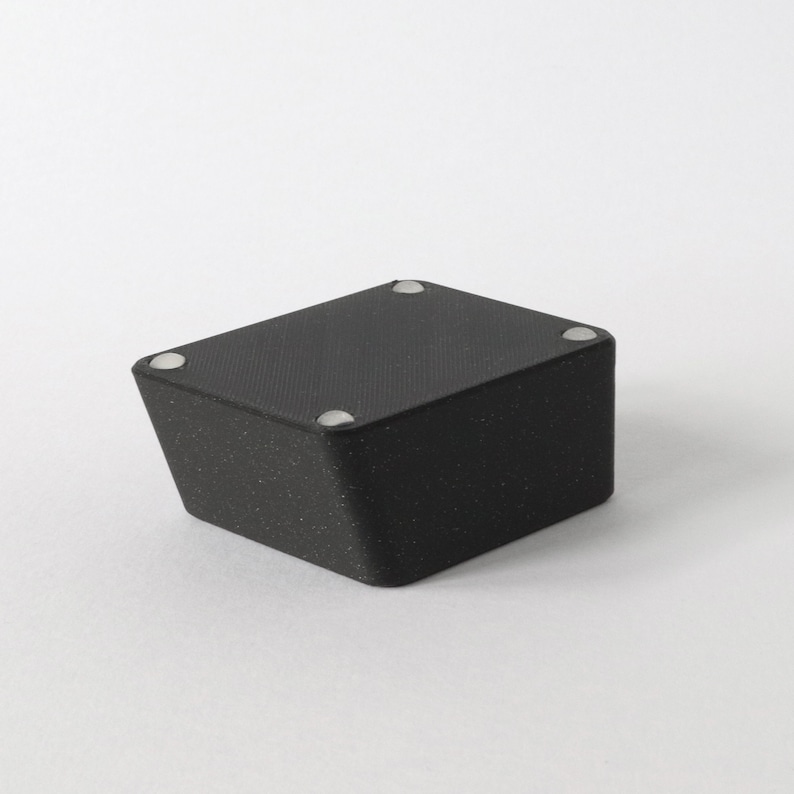 May include: Black square-shaped box with a textured surface and four silver dots on the top.