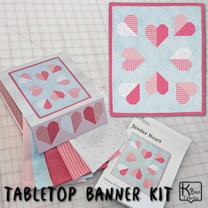 May include: A tabletop banner kit with a heart design. The kit includes a small quilt with pink and white hearts on a light blue background, fabric pieces, and a box with a matching heart pattern. The text "Tabletop Banner Kit" is visible.
