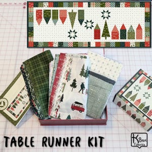 May include: A Christmas-themed table runner kit with a matching fabric bundle. The table runner features a festive design with pennants, stars, and houses. The fabric bundle includes various patterns with trees and a red van. The words "TABLE RUNNER KIT" are at the bottom.
