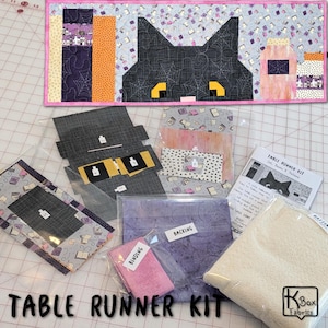 May include: A Halloween-themed table runner kit featuring a black cat design with yellow eyes and a pink border. The kit includes fabric pieces, binding, and backing. The words "TABLE RUNNER KIT" are printed at the bottom.