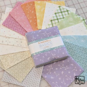 May include: A collection of pastel-colored fabric squares, including pink, orange, yellow, and green, arranged around a stack of lavender fabric squares. The stack is labeled "KIMBERBELL BASICS FLANNEL SWEET DREAMS 42 CHARMS". The fabrics feature floral and geometric patterns.