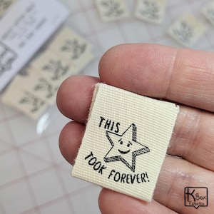 May include: Off-white fabric label with the text "THIS TOOK FOREVER!" and a smiling star graphic in black. The label is rectangular and appears to be for sewing or crafting projects. Several similar labels are visible in the background.