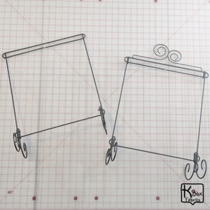 May include: Two black metal display stands with a decorative scroll design on the top of one stand. The stands are designed to hold fabric or other items.