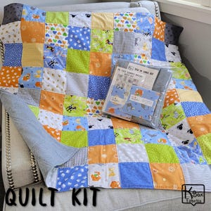 May include: A patchwork quilt featuring a grid of colorful squares with patterns of animals, hearts, and geometric shapes in blue, orange, green, and white. A clear plastic package containing a sewing label kit and fabric squares sits on top.